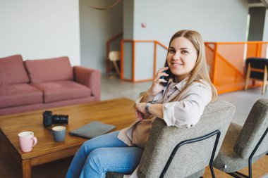 Bir kız öğrenci parlak bir kafede, bir kanepede oturuyor ve telefonda konuşuyor. Konforlu bir kafede uzaktan çalışma