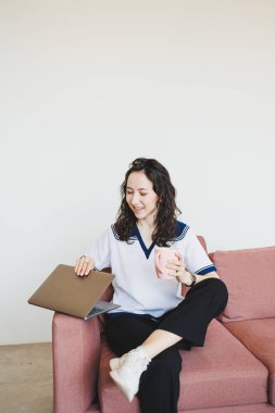 Parlak kafedeki kanepede oturan gülümseyen bayan öğrenci elinde kahveyle dizüstü bilgisayarına bakıyor. Samimi bir kafede dizüstü bilgisayarla uzaktan çalışma