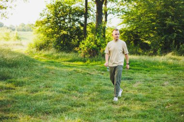 Bej tişörtlü ve pantolonlu yakışıklı genç bir adam yeşil çimlerde yürüyor. Şehirden uzakta doğada rahatlama.