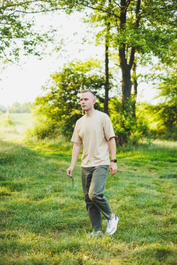 Bej tişörtlü ve pantolonlu yakışıklı genç bir adam yeşil çimlerde yürüyor. Şehirden uzakta doğada rahatlama.