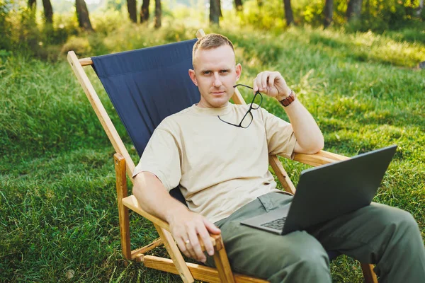 stock image A man works on a laptop while sitting on a bench in a city park. A young guy reads the news, writes a message on his laptop. Recreation in nature, remote work on vacation