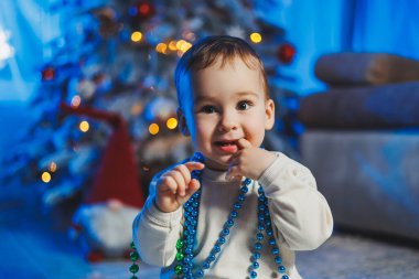 Ev ortamında bir Noel ağacının arka planında bej takım elbise giymiş küçük yakışıklı bir çocuk. Yeni yılı kutlamadan önce çocukların duyguları