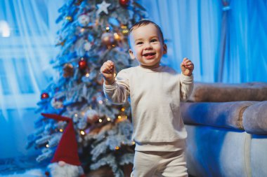 Ev ortamında bir Noel ağacının arka planında bej takım elbise giymiş küçük yakışıklı bir çocuk. Yeni yılı kutlamadan önce çocukların duyguları