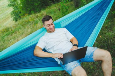 Dizüstü bilgisayarı olan genç bir adam hamakta oturuyor ve uzaktan çalışıyor. Tatil sırasında doğada çalışmak.