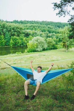 Dizüstü bilgisayarı olan genç bir adam hamakta oturuyor ve uzaktan çalışıyor. Tatil sırasında doğada çalışmak.