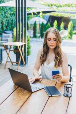 Genç bir kadın dizüstü bilgisayarda çalışıyor ve bir kafenin terasında rahat bir iş yerinde telefonla konuşuyor. Konforlu bir kafede uzaktan çalışma ya da tatillerde çalışma kavramı.