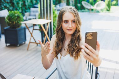 Genç bir kadın dizüstü bilgisayarda çalışıyor ve bir kafenin terasında rahat bir iş yerinde telefonla konuşuyor. Konforlu bir kafede uzaktan çalışma ya da tatillerde çalışma kavramı.