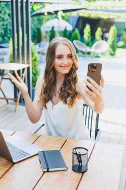 Genç bir kadın dizüstü bilgisayarda çalışıyor ve bir kafenin terasında rahat bir iş yerinde telefonla konuşuyor. Konforlu bir kafede uzaktan çalışma ya da tatillerde çalışma kavramı.