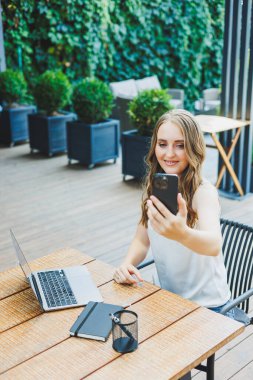 Genç bir kadın dizüstü bilgisayarda çalışıyor ve bir kafenin terasında rahat bir iş yerinde telefonla konuşuyor. Konforlu bir kafede uzaktan çalışma ya da tatillerde çalışma kavramı.