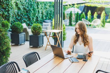 Genç bir kadın dizüstü bilgisayarda çalışıyor ve bir kafenin terasında rahat bir iş yerinde telefonla konuşuyor. Konforlu bir kafede uzaktan çalışma ya da tatillerde çalışma kavramı.