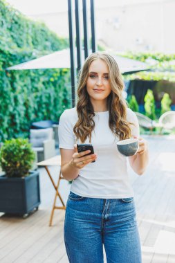 Serbest çalışan genç bir kadın, günlük giysiler içinde kahve içiyor ve telefonda konuşuyor, yeşil bir terasta çalışıyor, bir ekrana bakıyor ve bir kafede uzaktan bir proje üzerinde çalışıyor.