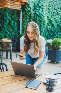Modern bir kafe caddesindeki bir kafede terasta oturan genç bir kadın. Telefonla masada oturan ve dışarıda dizüstü bilgisayar kullanan bir kadın. Bir yazlık kafede uzaktan çalışma