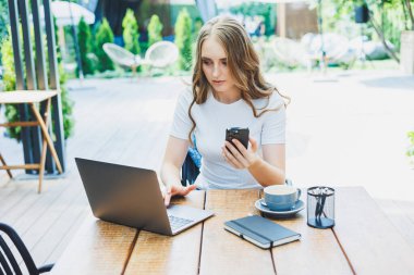 Modern bir kafe caddesindeki bir kafede terasta oturan genç bir kadın. Telefonla masada oturan ve dışarıda dizüstü bilgisayar kullanan bir kadın. Bir yazlık kafede uzaktan çalışma