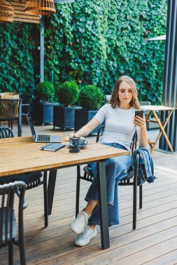 Modern bir kafe caddesindeki bir kafede terasta oturan genç bir kadın. Telefonla masada oturan ve dışarıda dizüstü bilgisayar kullanan bir kadın. Bir yazlık kafede uzaktan çalışma
