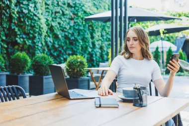 Modern bir kafe caddesindeki bir kafede terasta oturan genç bir kadın. Telefonla masada oturan ve dışarıda dizüstü bilgisayar kullanan bir kadın. Bir yazlık kafede uzaktan çalışma