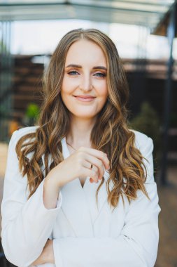 Yüzünde bir gülümseme olan beyaz takım elbiseli şık bir kadın bir yaz kafesinin terasında duruyor..