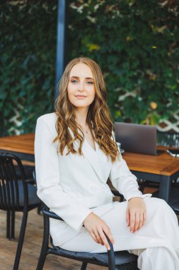 Yüzünde bir gülümseme olan beyaz takım elbiseli şık bir kadın bir yaz kafesinin terasında duruyor..