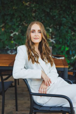 Yüzünde bir gülümseme olan beyaz takım elbiseli şık bir kadın bir yaz kafesinin terasında duruyor..