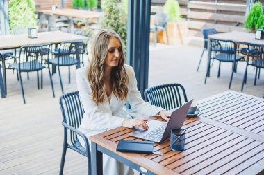 Beyaz, klasik takım elbiseli güzel bir kadın bir kafedeki terasta dizüstü bilgisayarla çalışıyor. Şık iş kıyafetleri içinde bir kadın bir yazlık kafede dizüstü bilgisayarla uzaktan çalışıyor.