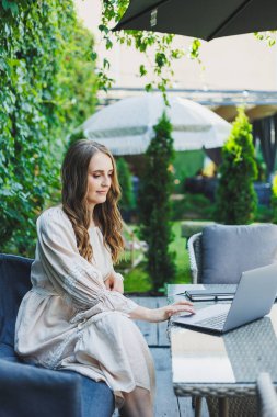 Yazlık terasta dizüstü bilgisayarda bağımsız çalışan bir kadın. Elbise giymiş bir iş kadını su içer ve dizüstü bilgisayarda çalışır.