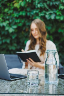 Yazlık terasta dizüstü bilgisayarda bağımsız çalışan bir kadın. Elbise giymiş bir iş kadını su içer ve dizüstü bilgisayarda çalışır.