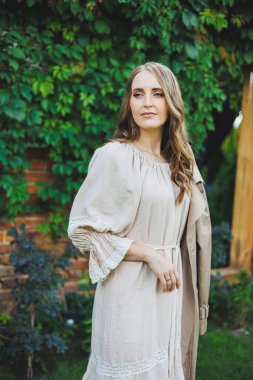 Açık bej elbiseli güzel bir genç kadının portresi. Yeşil bir bahçenin arka planında yüzünde gülümseme olan elbiseli romantik bir kadın. Bayram için kadın kokteyl elbisesi.