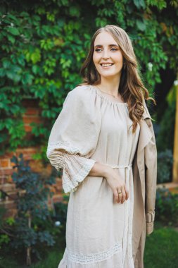 Açık bej elbiseli güzel bir genç kadının portresi. Yeşil bir bahçenin arka planında yüzünde gülümseme olan elbiseli romantik bir kadın. Bayram için kadın kokteyl elbisesi.