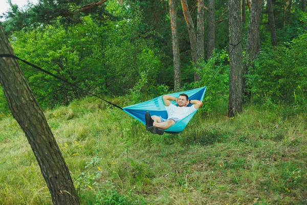 A young man rests in nature sitting in a tourist hammock. Hammock for outdoor recreation. Weekend rest of a lonely man in a deserted forest