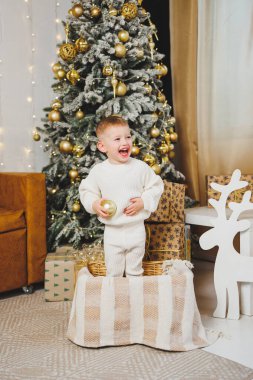 2-3 yaşlarında neşeli bir çocuk Noel ağacının yanında duruyor etrafı çelenkler ve Noel hediyeleriyle çevrili. Noel masalı konsepti, küçük çocuklar için bir tatil. Noel ağacı