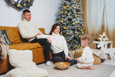 Küçük çocuklu neşeli bir genç aile evdeki Noel ağacının yanında dinleniyor. Evde Noel süsü olan aile çifti. Noel, Yeni Yıl - kutlama zamanı