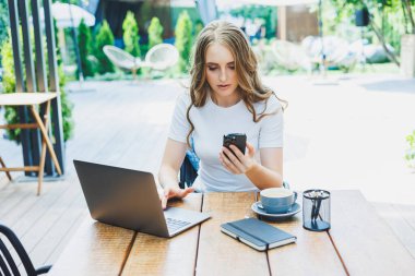 Modern bir kafe caddesindeki bir kafede terasta oturan genç bir kadın. Telefonla masada oturan ve dışarıda dizüstü bilgisayar kullanan bir kadın. Bir yazlık kafede uzaktan çalışma
