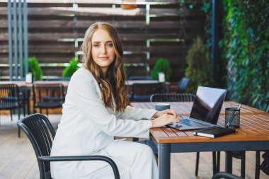 Beyaz, klasik takım elbiseli güzel bir kadın bir kafedeki terasta dizüstü bilgisayarla çalışıyor. Şık iş kıyafetleri içinde bir kadın bir yazlık kafede dizüstü bilgisayarla uzaktan çalışıyor.