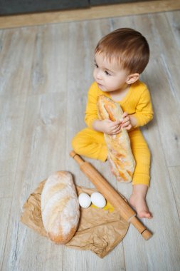 1 yaşında tatlı bir çocuk mutfakta oturuyor ve taze ekmek yiyor. Yerde ekmeği olan çocuk.