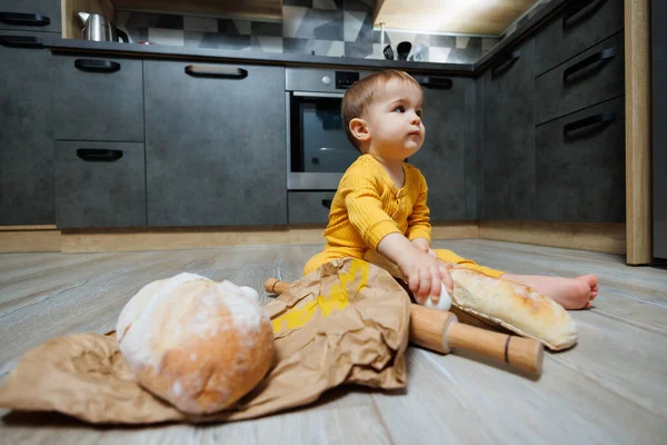 A cute 1-year-old boy is sitting in the kitchen with fresh bread. Child with bread on the floor. The child eats bread