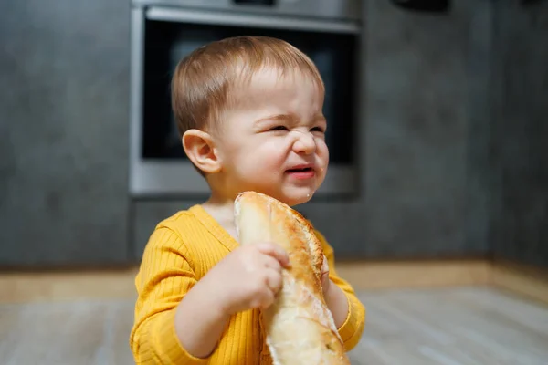 A cute 1-year-old boy is sitting in the kitchen with fresh bread. Child with bread on the floor. The child eats bread