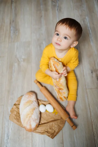 A cute 1-year-old boy is sitting in the kitchen and eating fresh bread. Child with bread on the floor.