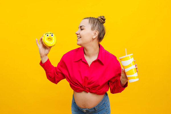 A smiling pregnant woman in a pink shirt is eating a sweet cake and drinking a sweet drink. Happy pregnant woman eats unhealthy sweet donuts.