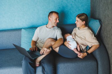 Mutlu aile anne, baba ve tatlı bebek evde dizüstü bilgisayar kullanırken eğleniyorlar. Genç ebeveynler ve küçük çocuk bilgisayar başında kanepede oturarak zaman geçiriyorlar.