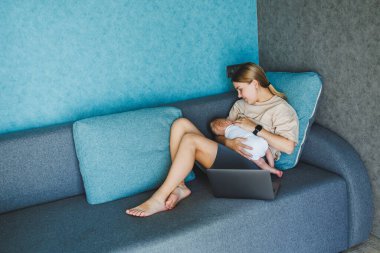 Gülümseyen genç bir anne bebeğini emziriyor ve e-posta yazarken bebeğiyle birlikte kanepesinde çalışıyor. Bebeği emziriyorum. Evde çalışan genç bir kadın..