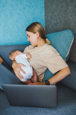 Gülümseyen genç bir anne bebeğini emziriyor ve e-posta yazarken bebeğiyle birlikte kanepesinde çalışıyor. Bebeği emziriyorum. Evde çalışan genç bir kadın..