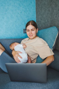 Gülümseyen genç bir anne bebeğini emziriyor ve e-posta yazarken bebeğiyle birlikte kanepesinde çalışıyor. Bebeği emziriyorum. Evde çalışan genç bir kadın..