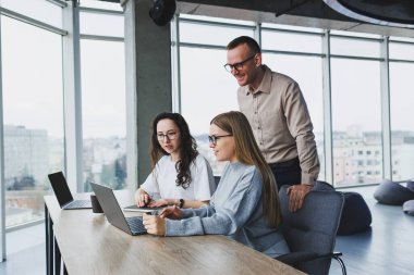 Modern bir ofiste üç çalışan, bir adam ve bir kadın masada çalışıyor, iş arkadaşları ortak bir proje hakkında tartışıyor ve fikir alışverişinde bulunuyor..
