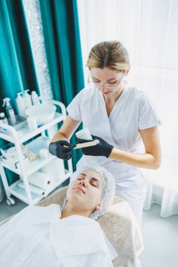 Masaj masasında oturan, genç bir kadının yüzüne masaj yapan ve yüz bakımı yapan kadın hastalarla birlikte çalışan bir dermatolog ya da kozmetikçinin ofisi. Yüz bakımı.