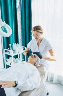 Masaj masasında oturan, genç bir kadının yüzüne masaj yapan ve yüz bakımı yapan kadın hastalarla birlikte çalışan bir dermatolog ya da kozmetikçinin ofisi. Yüz bakımı.