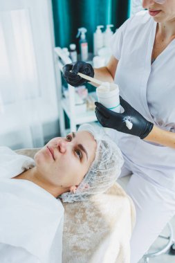 Masaj masasında oturan, genç bir kadının yüzüne masaj yapan ve yüz bakımı yapan kadın hastalarla birlikte çalışan bir dermatolog ya da kozmetikçinin ofisi. Yüz bakımı.