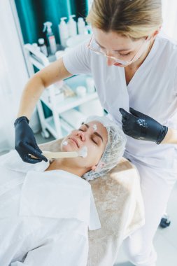 Masaj masasında oturan, genç bir kadının yüzüne masaj yapan ve yüz bakımı yapan kadın hastalarla birlikte çalışan bir dermatolog ya da kozmetikçinin ofisi. Yüz bakımı.