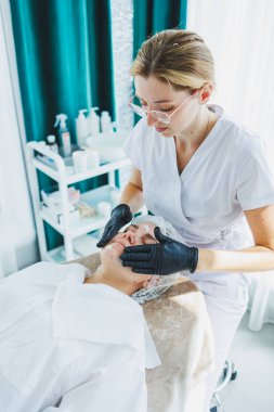 Masaj masasında oturan, genç bir kadının yüzüne masaj yapan ve yüz bakımı yapan kadın hastalarla birlikte çalışan bir dermatolog ya da kozmetikçinin ofisi. Yüz bakımı.