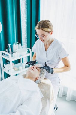 Masaj masasında oturan, genç bir kadının yüzüne masaj yapan ve yüz bakımı yapan kadın hastalarla birlikte çalışan bir dermatolog ya da kozmetikçinin ofisi. Yüz bakımı.