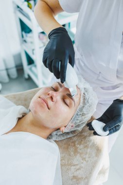 Klinikteki bir cildiyeci ya da kozmetikçi kadın hastalarla çalışır ve genç bir kadının yüzünde yüz bakımı yapar. Kozmetik uzmanının ofisindeki yüz ultrasonik temizleme.