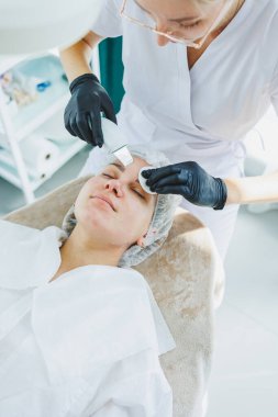 Klinikteki bir cildiyeci ya da kozmetikçi kadın hastalarla çalışır ve genç bir kadının yüzünde yüz bakımı yapar. Kozmetik uzmanının ofisindeki yüz ultrasonik temizleme.